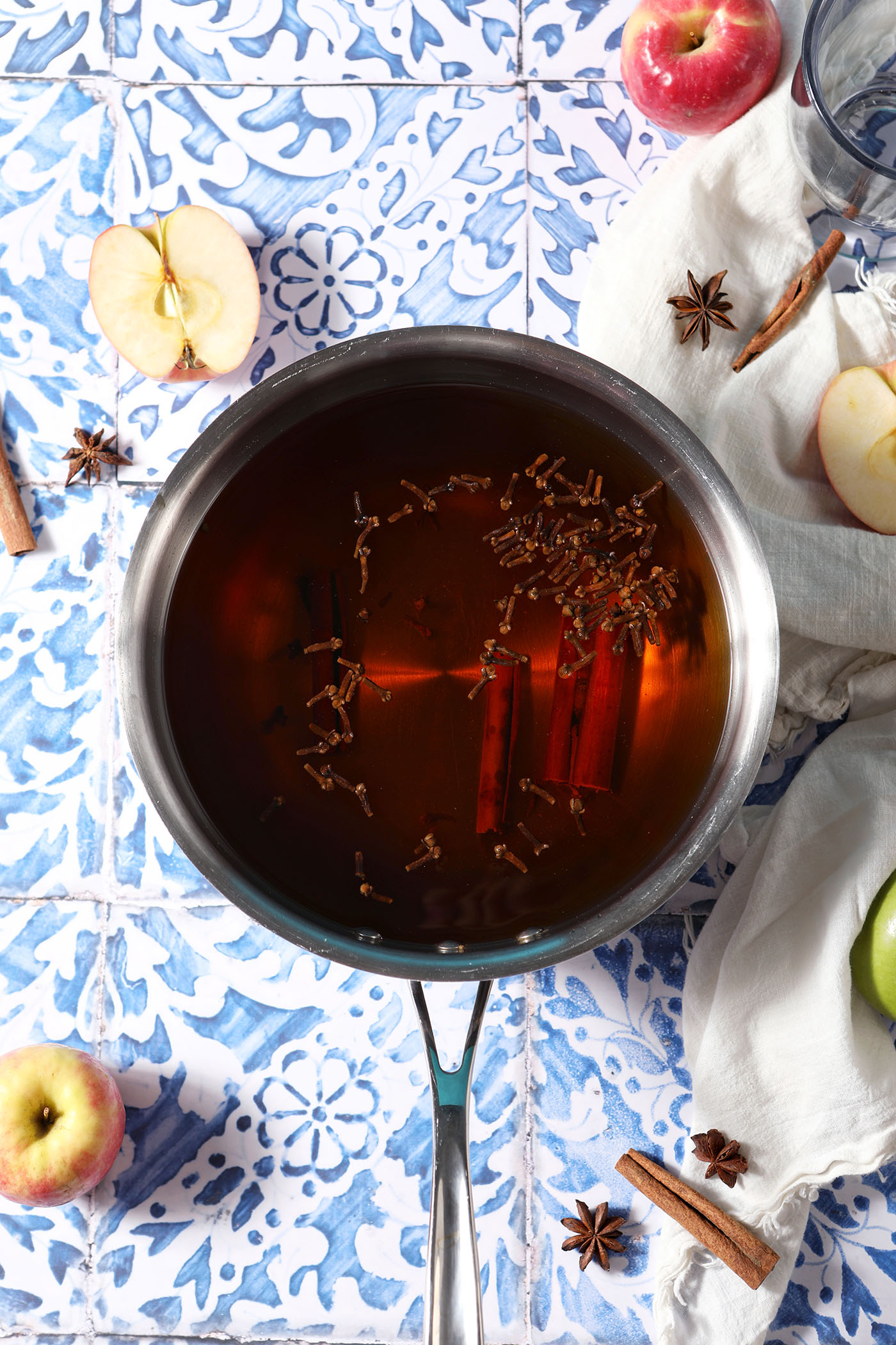 Overhead of a saucepan holding spiced apple cider with spices in it on a blue tiled surface surrounded by apples and whole spices