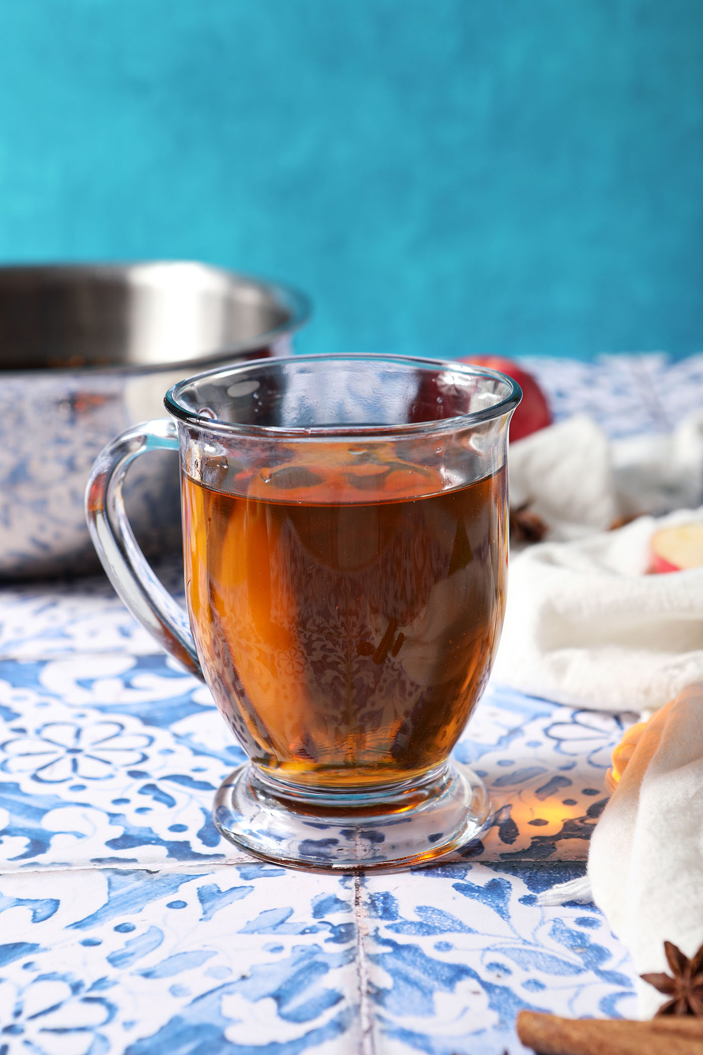 A mug of easy spiced cider on a blue tiled surface in front of a saucepan