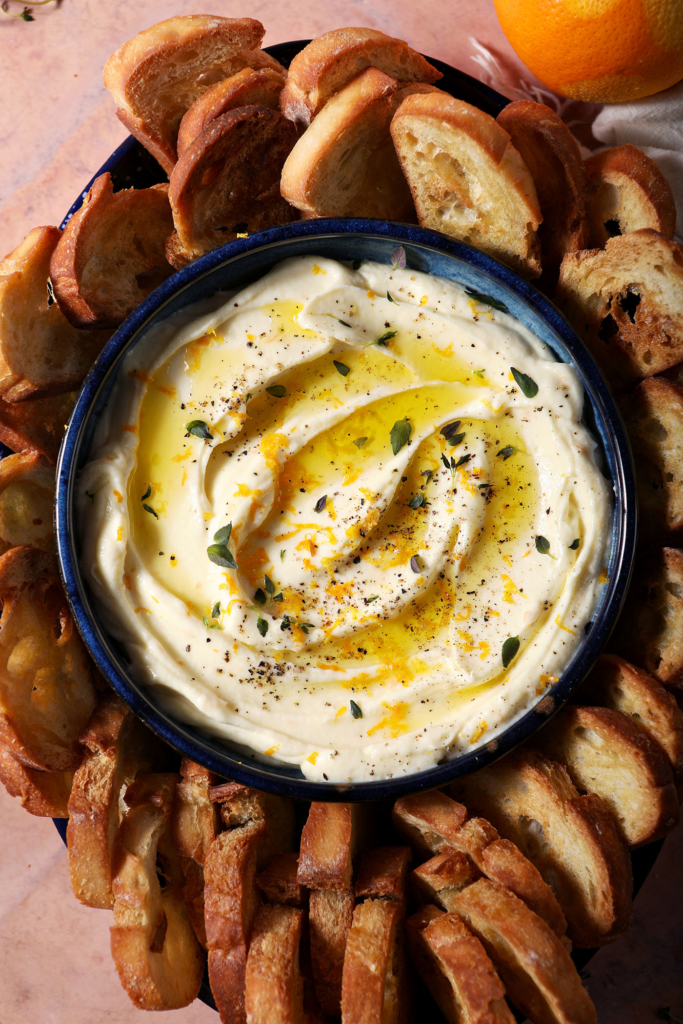 A bowl of whipped ricotta dip garnished with extra thyme, orange zest and olive oil surrounded by crostini on a terracotta surface