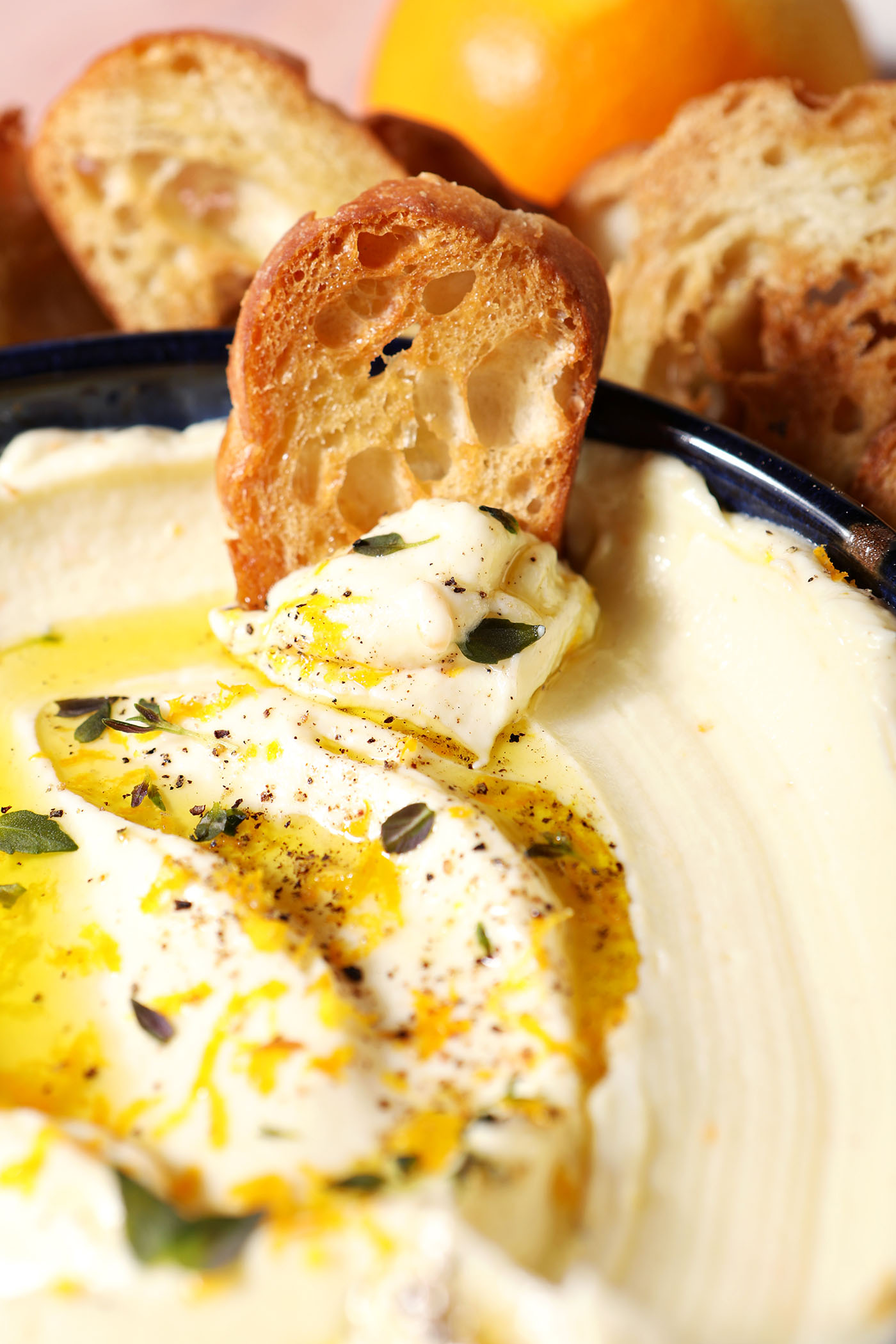 A crostini sits on top of a bowl of whipped ricotta garnished with oil, herbs and orange zest