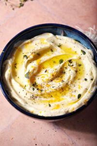 A bowl of whipped ricotta dip garnished with oil, orange zest and fresh herbs on a terracotta surface