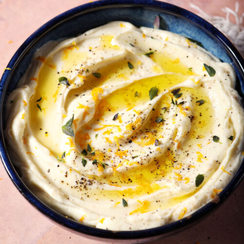 A bowl of whipped ricotta dip garnished with oil, orange zest and fresh herbs on a terracotta surface