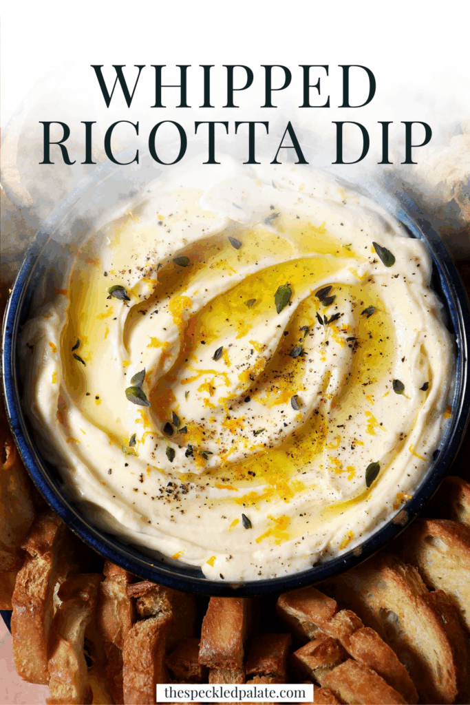 A bowl of whipped ricotta dip garnished with extra thyme, orange zest and olive oil surrounded by crostini on a terracotta surface with the text whipped ricotta dip