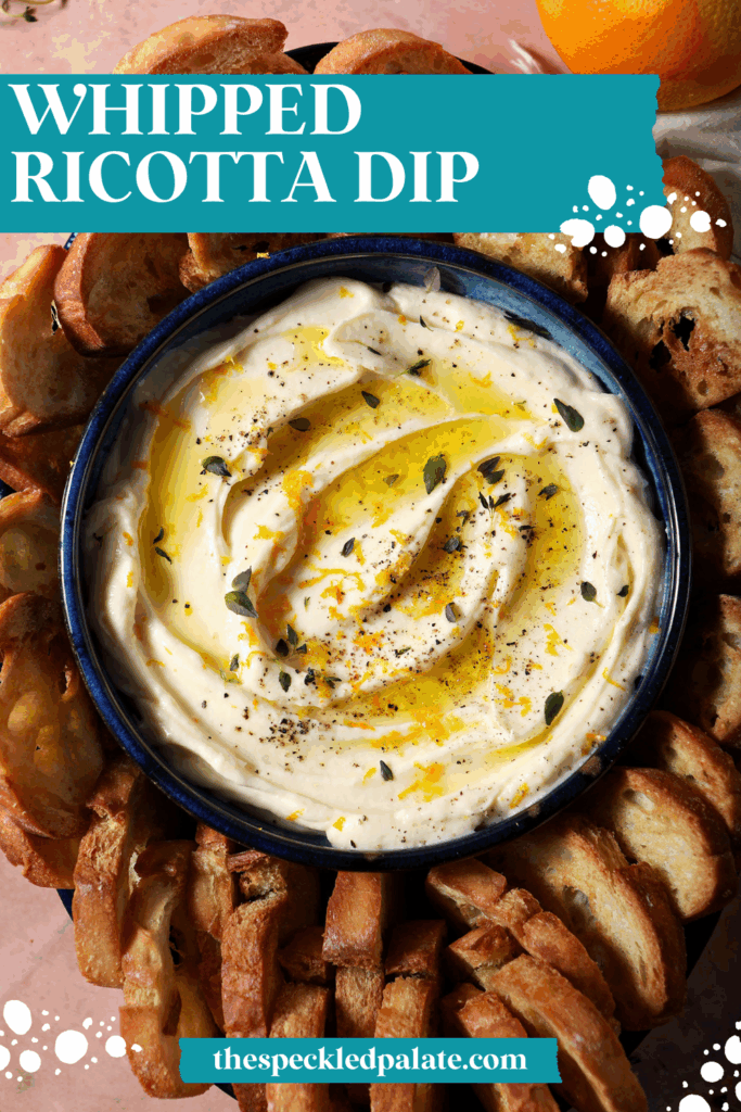 A bowl of whipped ricotta dip garnished with extra thyme, orange zest and olive oil surrounded by crostini on a terracotta surface with the text whipped ricotta dip