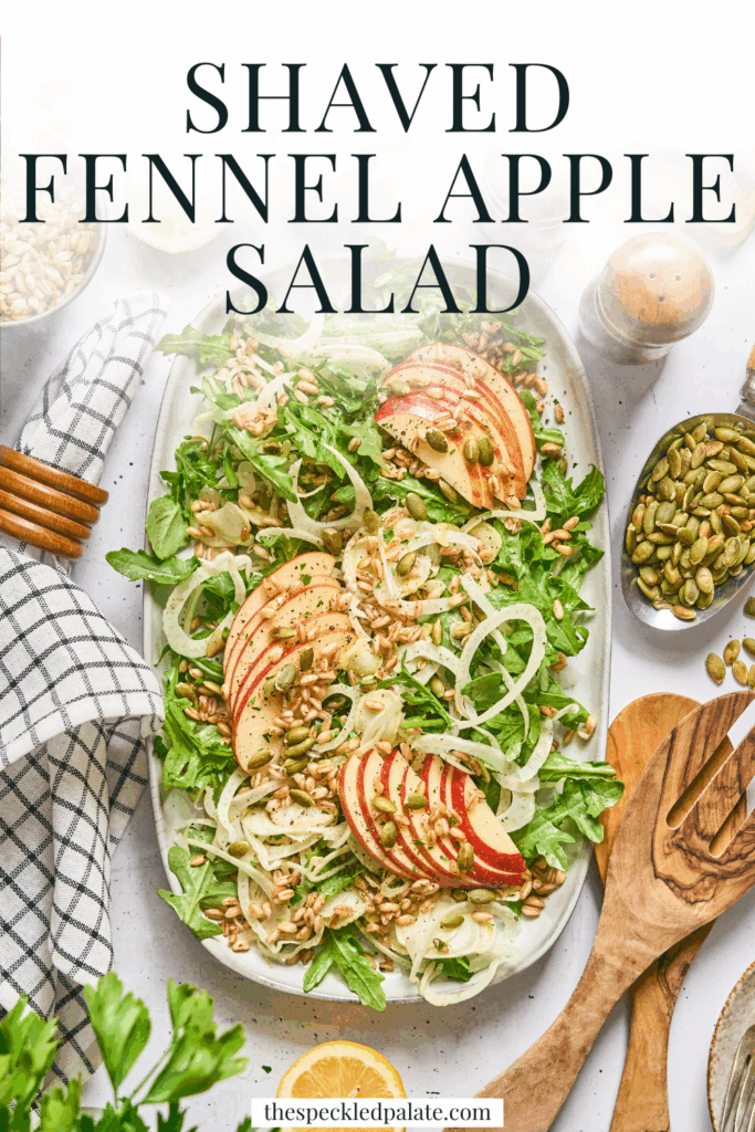 Overhead of a large serving plate of Shaved Fennel and Apple Farro Salad, surrounded by ingredients, wooden utensils and a blue patterned linen with the text shaved fennel apple salad