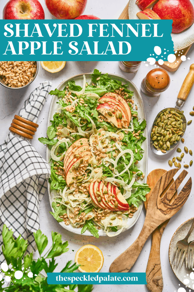 Overhead of a large serving plate of Shaved Fennel and Apple Farro Salad, surrounded by ingredients, wooden utensils and a blue patterned linen with the text shaved fennel apple salad
