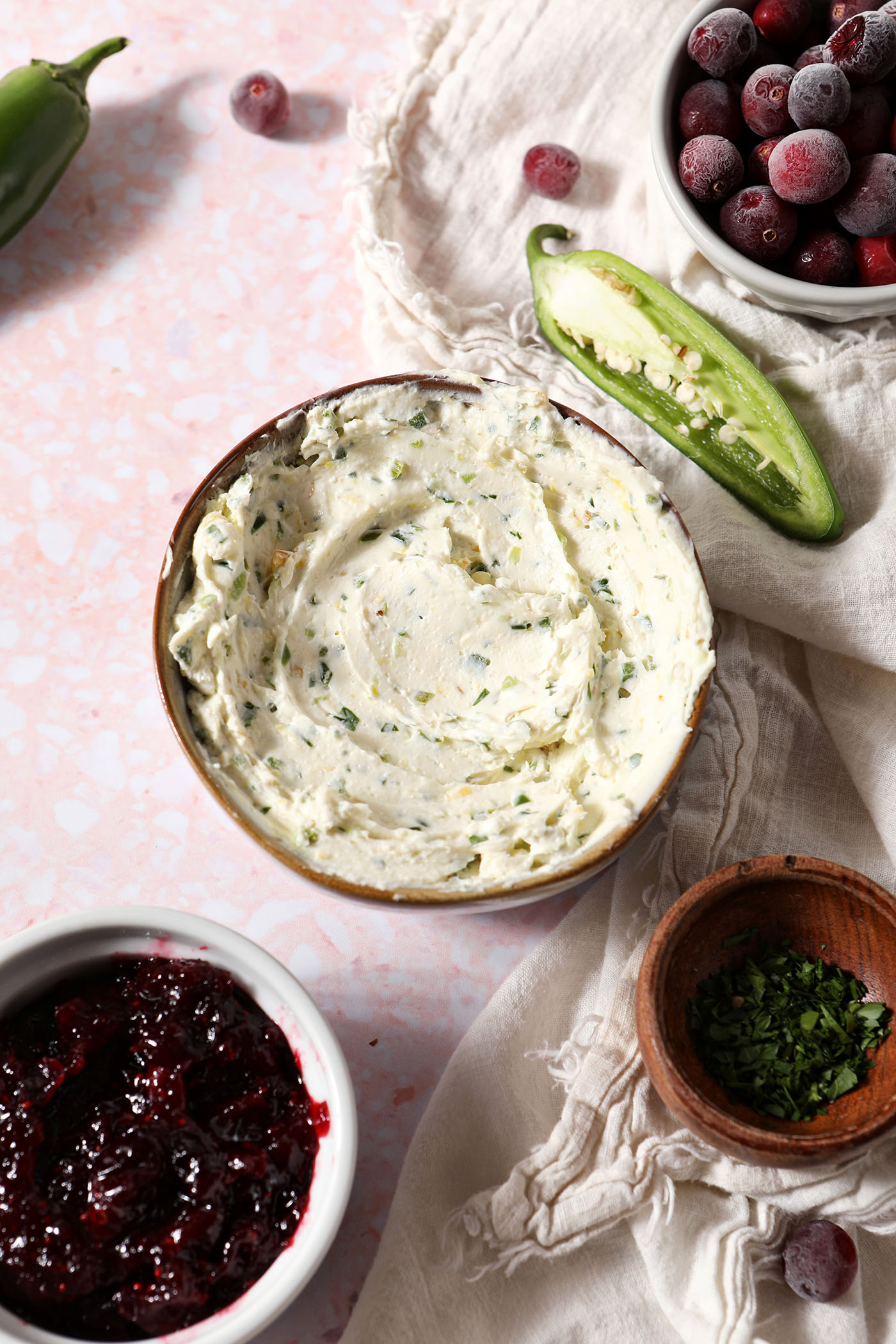 Bowls hold jalapeño cream cheese, cranberry sauce and chopped parsley on a pink surface surrounded by cranberries and jalapeño peppers