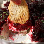A piece of bread dips into a bowl of creamy cranberry dip