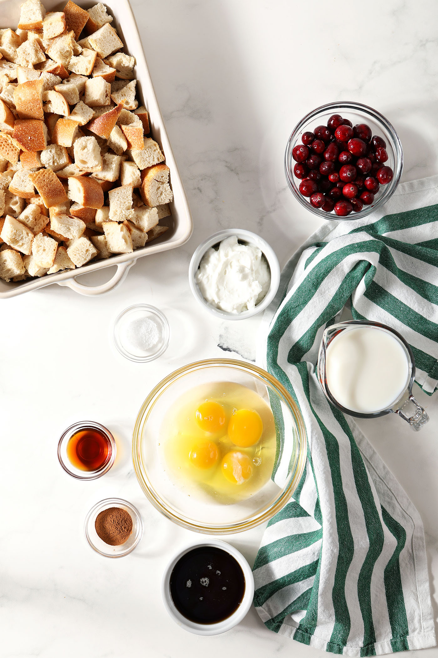 Ingredients to make overnight French casserole with cranberries in bowls on a white stone surface next to a green and white striped linen