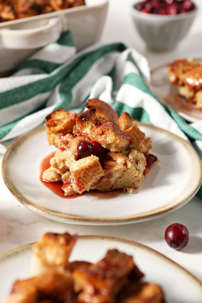 A plate of French toast casserole with cranberries on a plate next to other plates and the casserole dish with a green and white striped linen