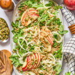Overhead of a large serving plate of Shaved Fennel and Apple Farro Salad, surrounded by ingredients, wooden utensils and a blue patterned linen
