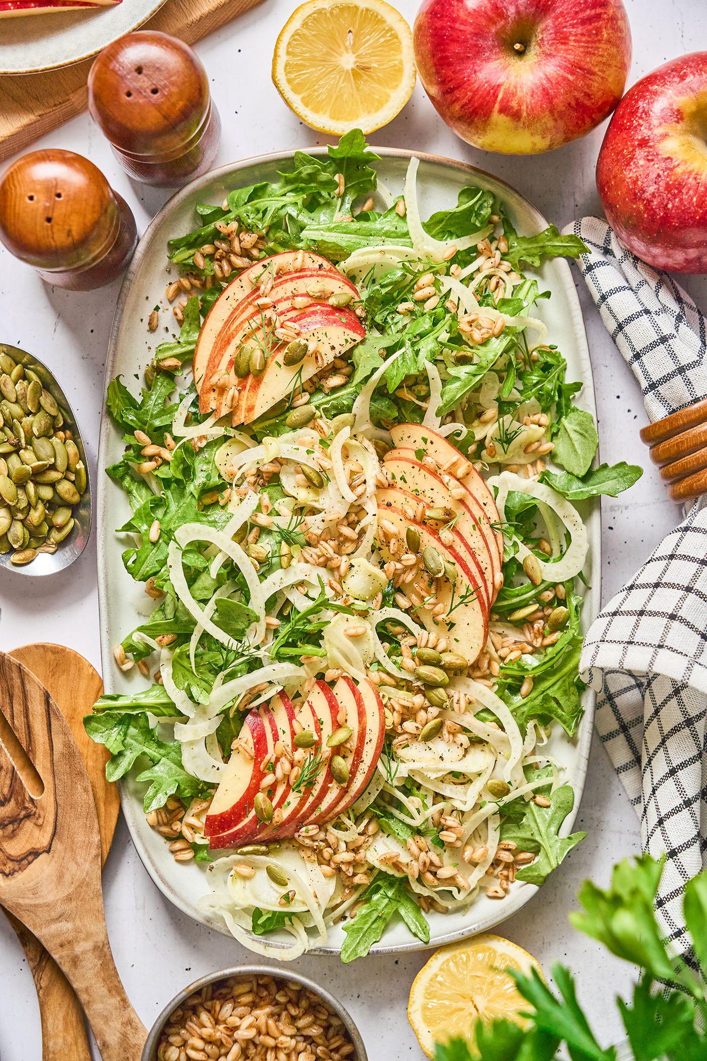 Overhead of a large serving plate of Shaved Fennel and Apple Farro Salad, surrounded by ingredients, wooden utensils and a blue patterned linen