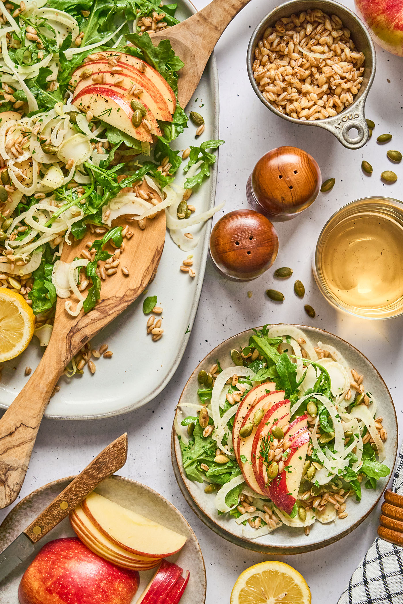 A platter of salad sits next to a plate holding the salad, topped with sliced apples, and surrounded on the surface with various ingredients