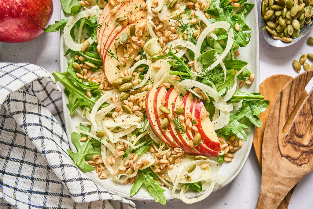 Close up of a platter holding a beautifully styled fennel apple salad with farro next to a blue checked linen and wooden utensils