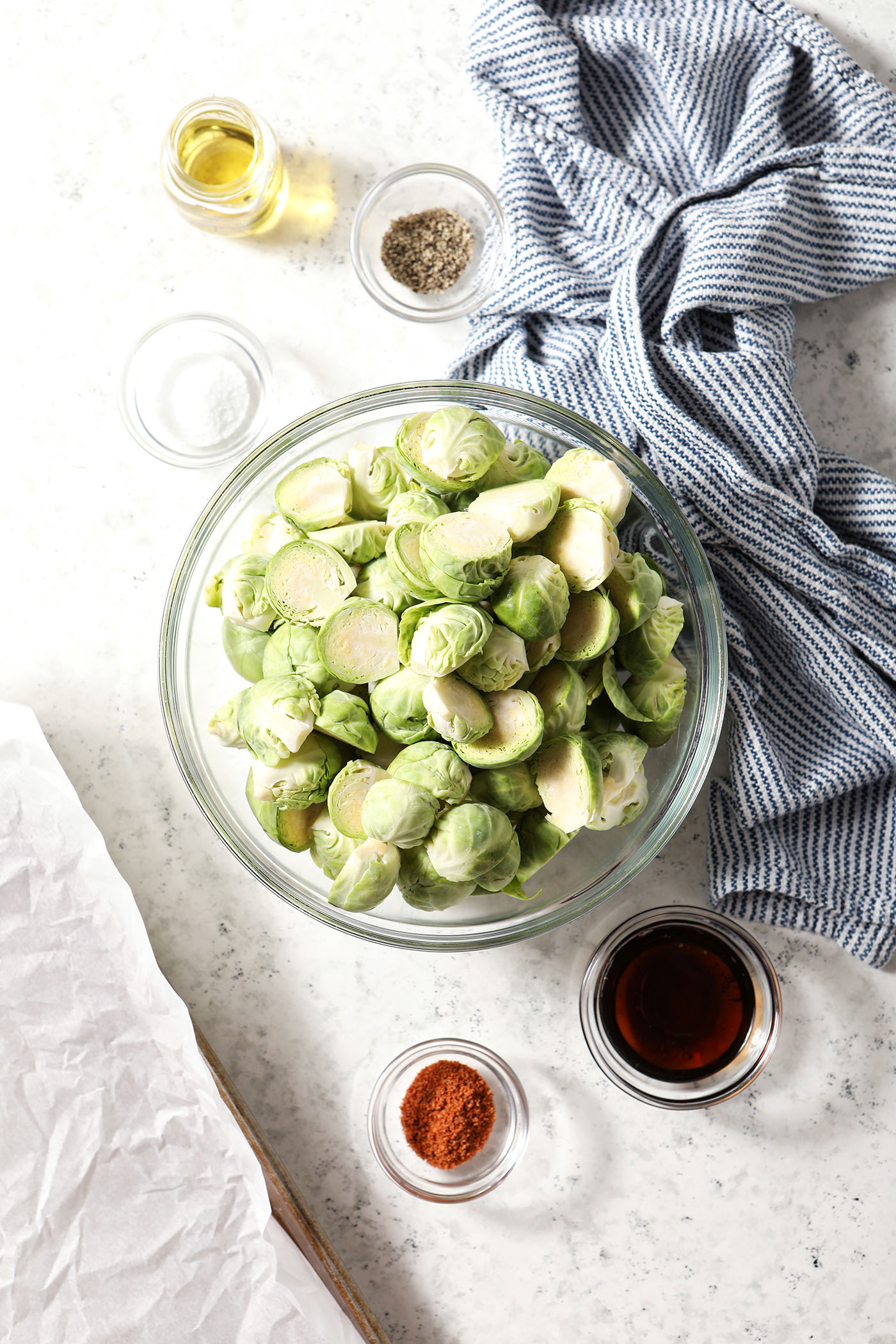A large bowl of halved Brussels sprouts surrounded by other ingredients to make roasted Brussels sprouts on a white stone surface with a blue striped linen