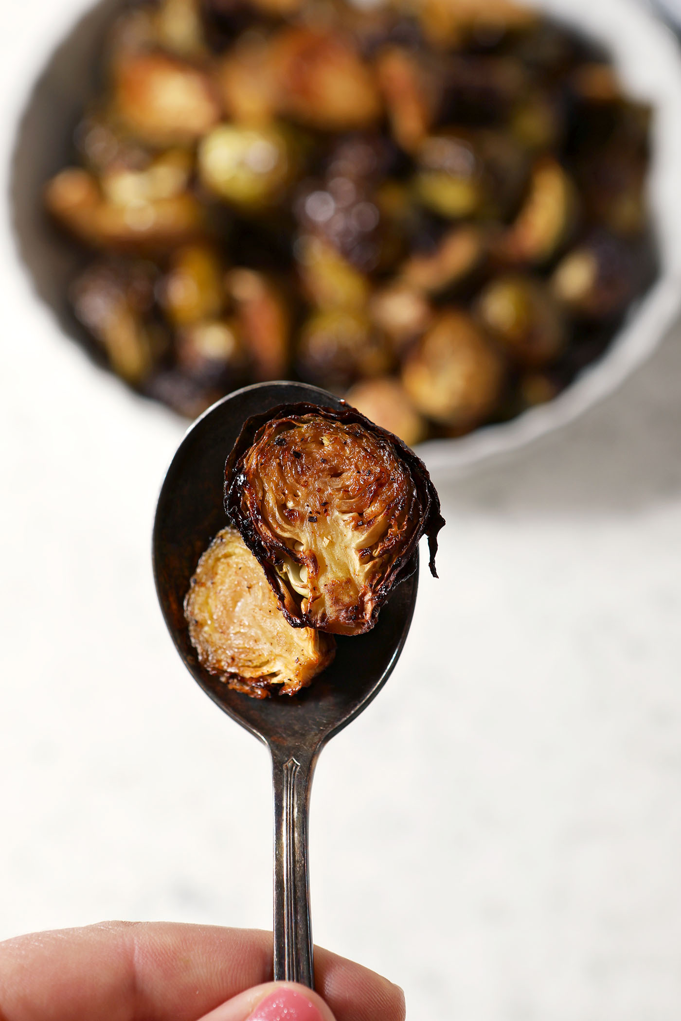 A spoon holds two halved roasted brussels sprouts with maple above a stone surface and a bowl of more sprouts