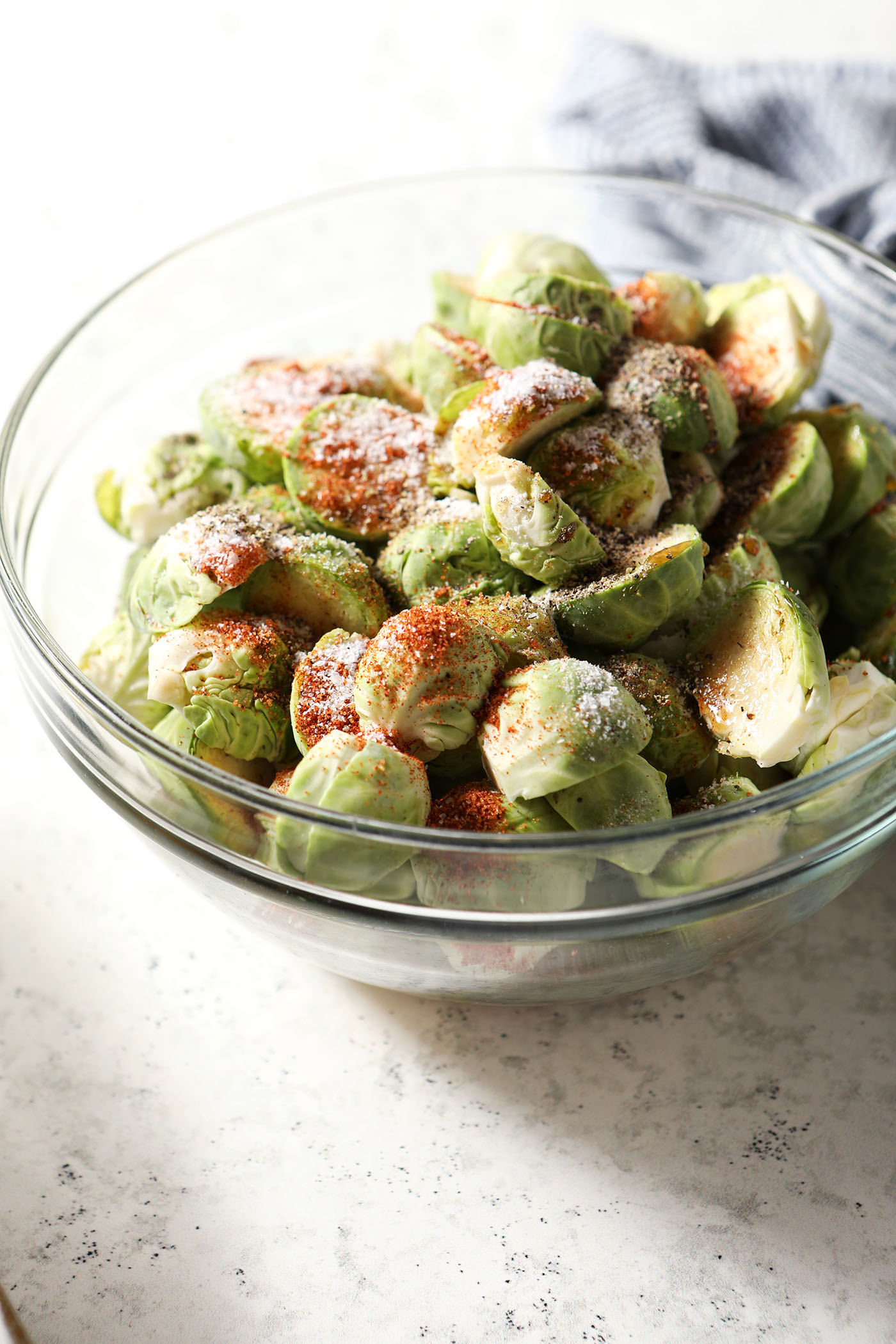 A bowl holds halved brussels sprouts with seasonings on top of them, before tossing, on a white stone surface