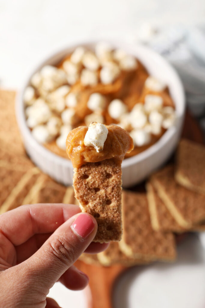 A hand holds a graham cracker with a bite of sweet potato pie dip on it
