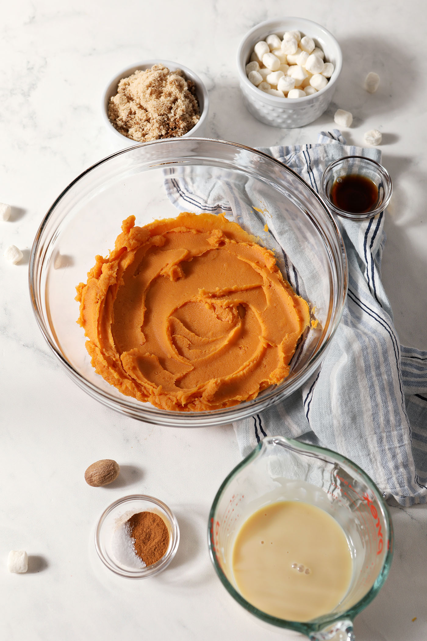 Ingredients to make Sweet Potato Pie Dip in bowls and containers on a white stone surface next to a blue and white striped linen