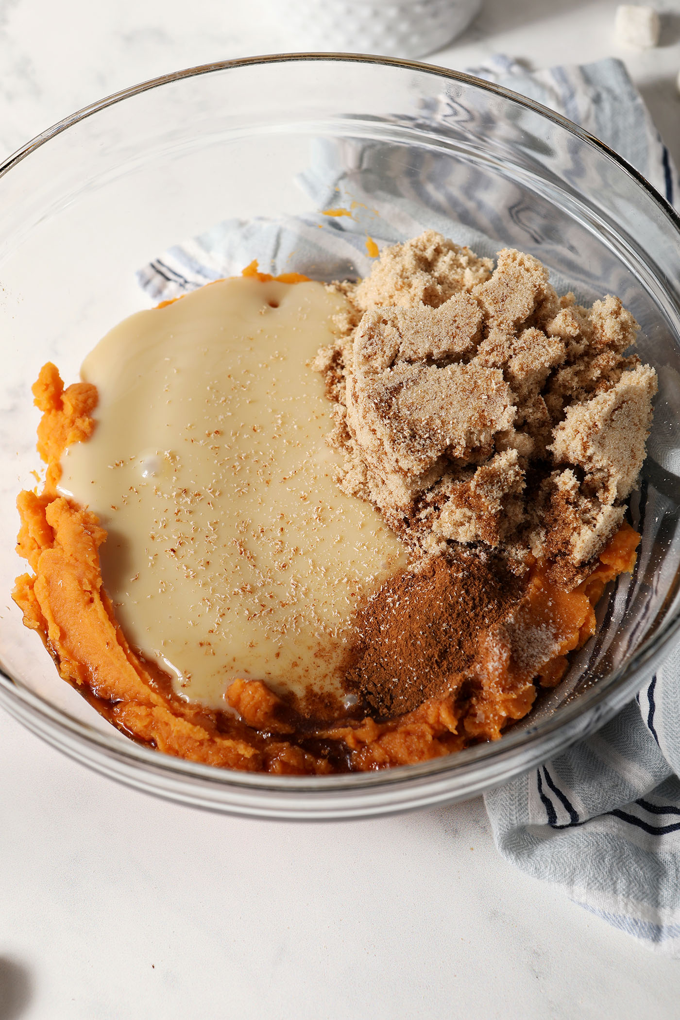 Ingredients to make sweet potato dessert dip in a glass bowl on a white stone surface next to a blue and white striped linen