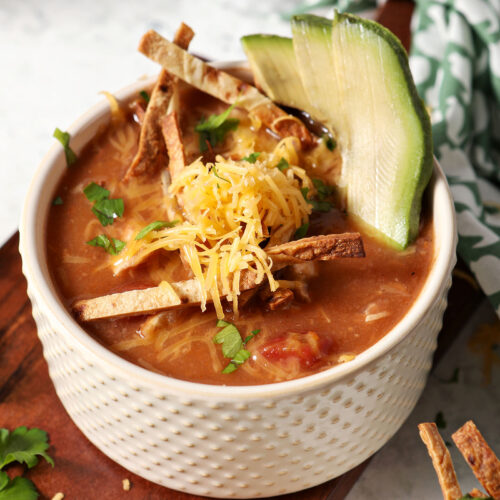 A bowl of turkey tortilla soup garnished with tortilla slivers, avocado, cilantro and grated cheese sits on top of a wooden slab