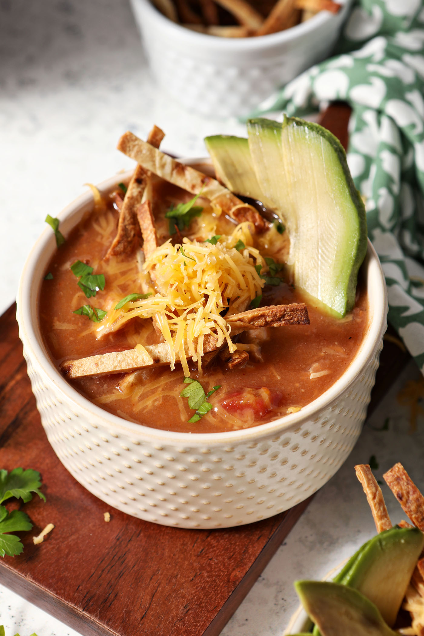 A bowl of turkey tortilla soup garnished with tortilla slivers, avocado, cilantro and grated cheese sits on top of a wooden slab