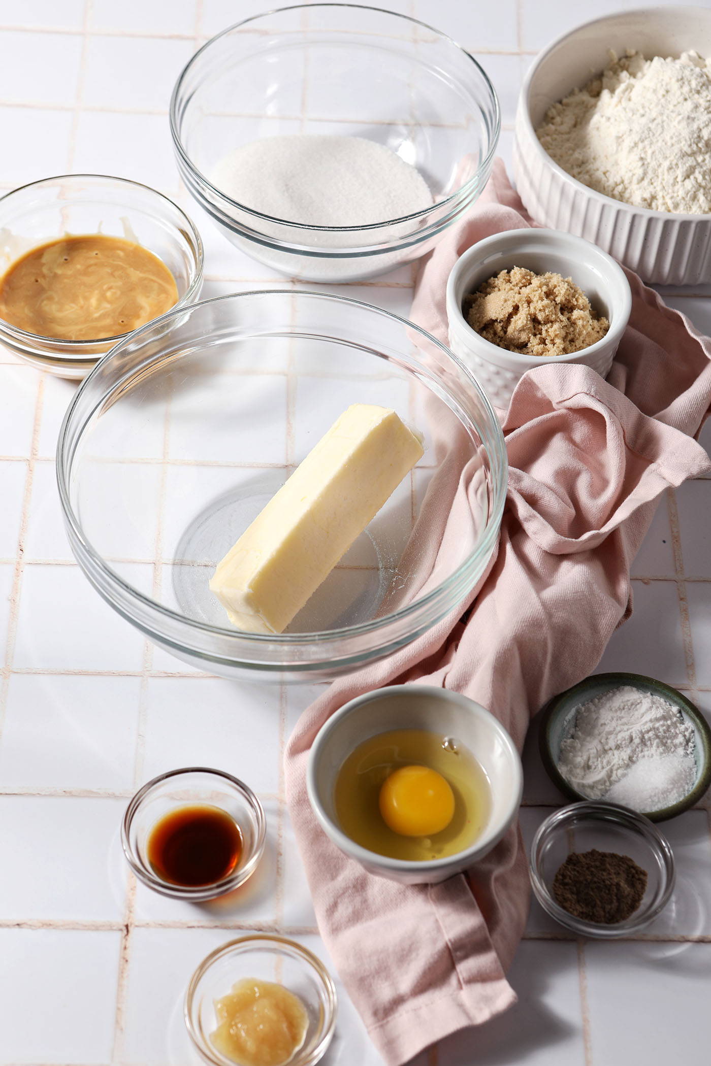 Ingredients to make spiced maple cookies in bowls on a white tiled surface next to a pink linen
