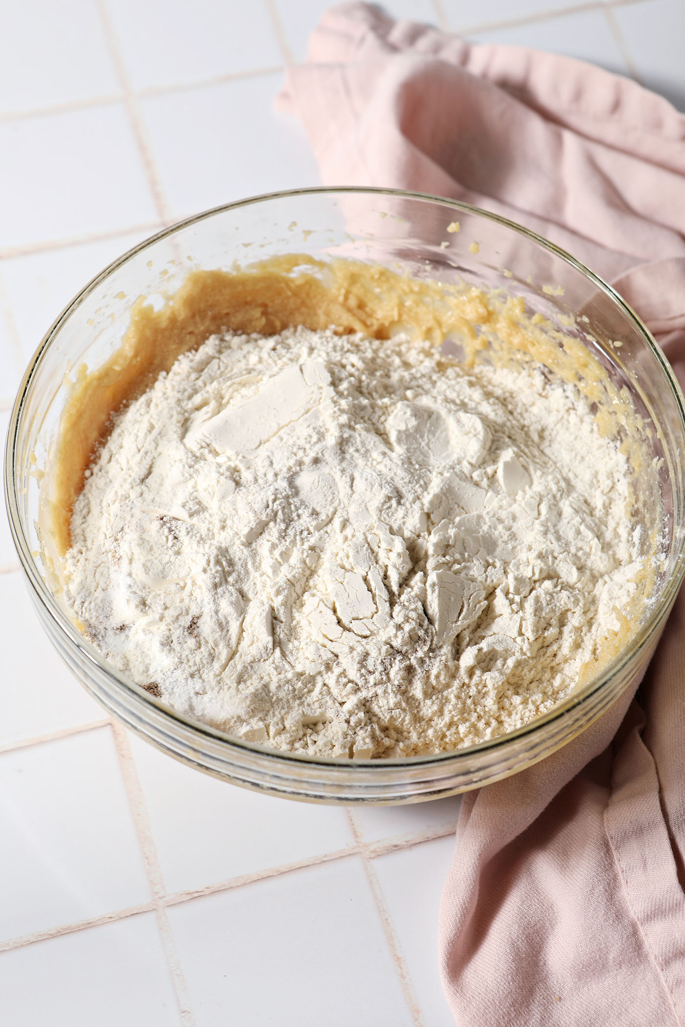 Dry ingredients sit on top of wet ingredients in a bowl on a white tiled surface next to a pink linen