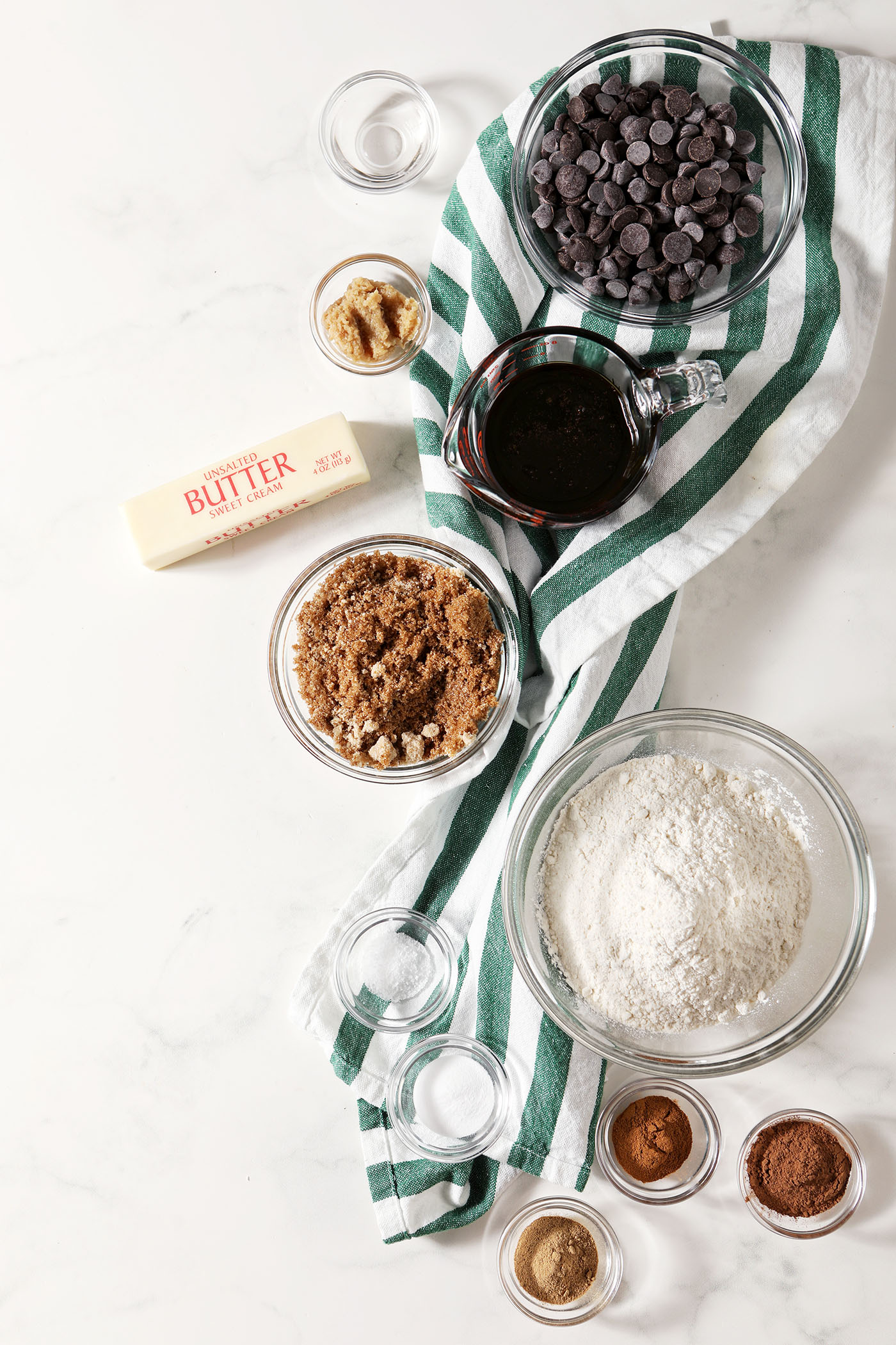 Ingredients to make chocolate ginger cookies in bowls with a green and white striped linen on a white stone surface
