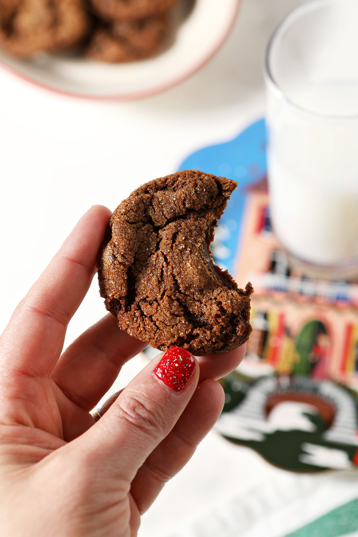 A hand holds a bitten-into chocolate ginger cookie in front of a holiday plate with a glass of milk on it