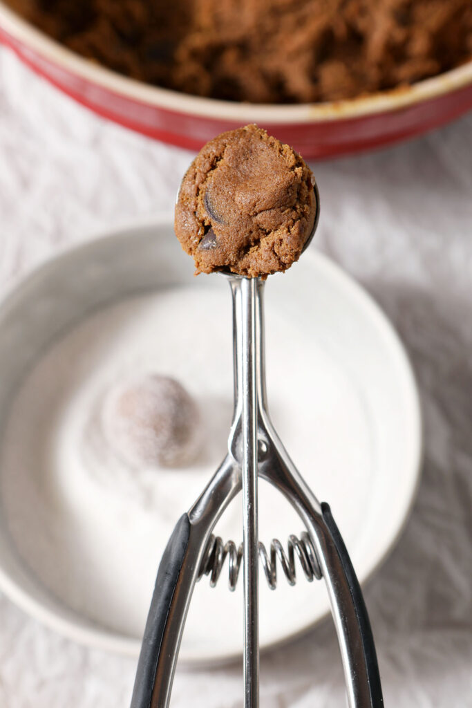 A cookie scoop holds a scoop of a cookie above a sheet pan holding a bowl of sugar