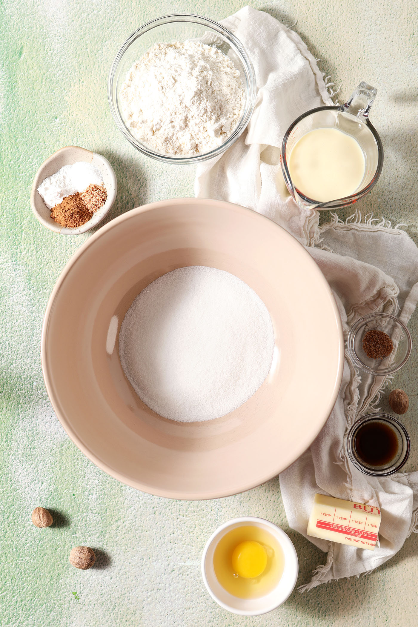 Ingredients to make cookies with eggnog in bowls on a green surface next to a beige linen