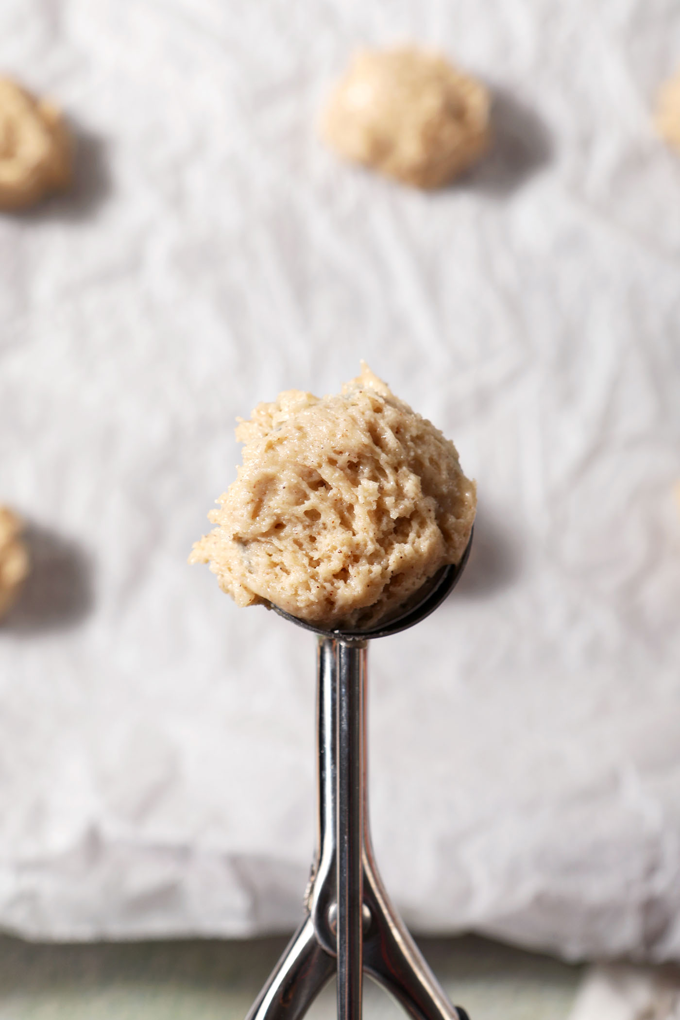 A cookie scoop holds a scoop of eggnog cookie batter above a parchment-lined sheet pan