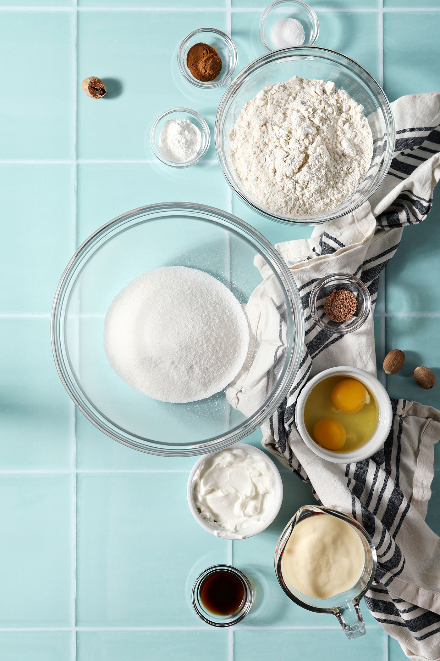 Ingredients to make muffins with eggnog in bowls on a green tiled surface next to a black and white linen