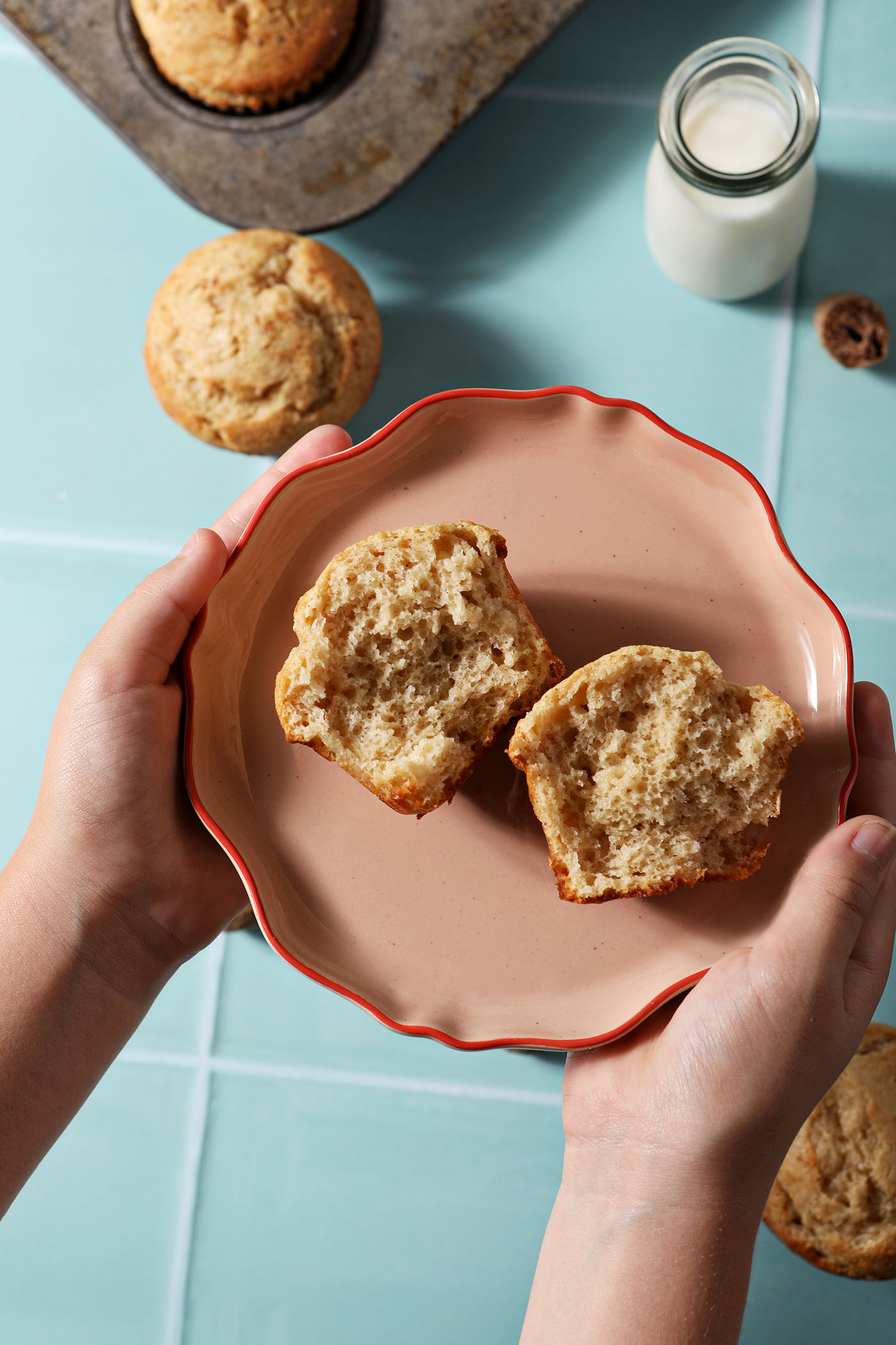 A child holds a pink plate with a halved eggnog muffin on top of it above a green tiled surface with more muffins