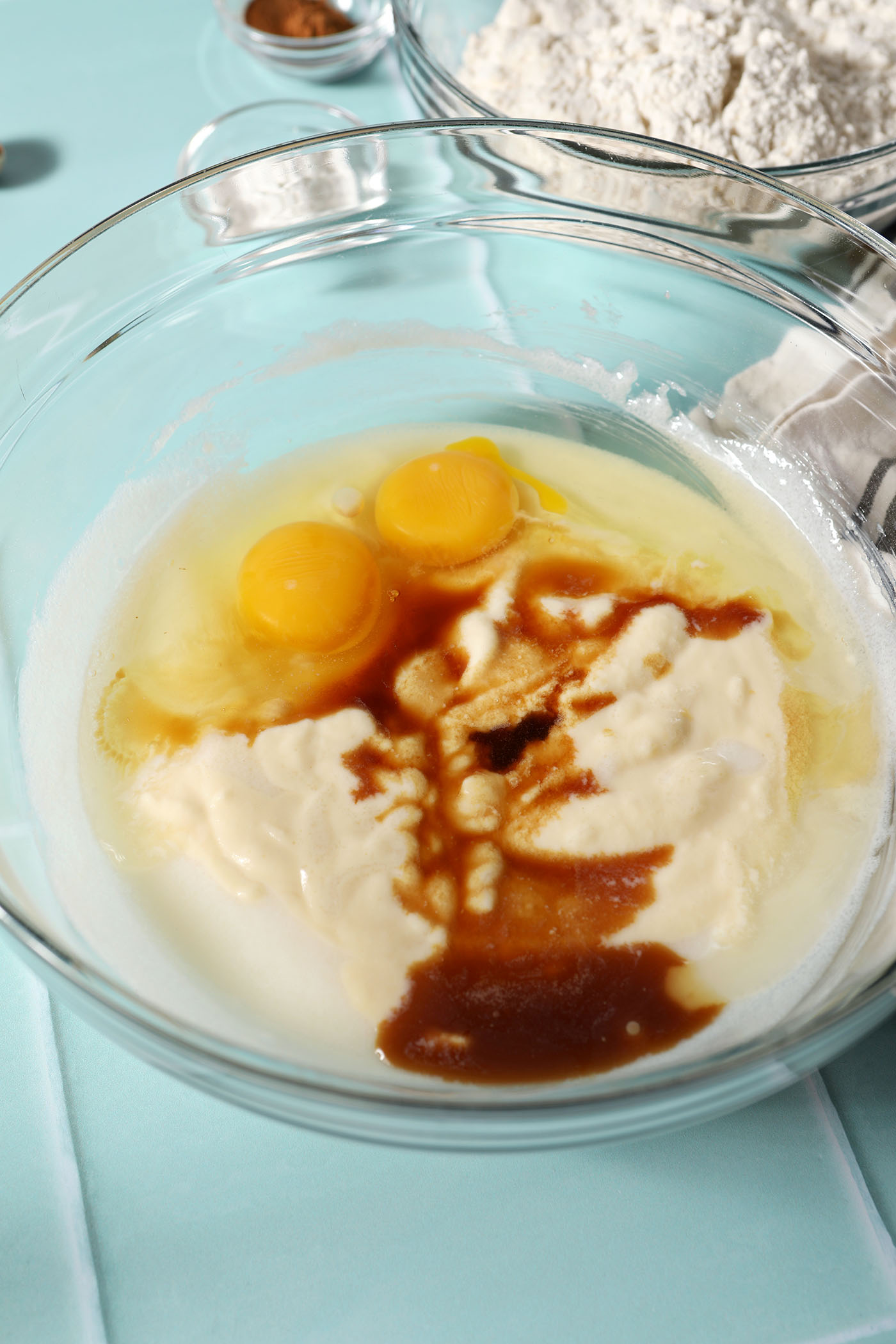 Eggs, eggnog and vanilla extract on top of other wet ingredients in a bowl on a green tiled surface before mixing
