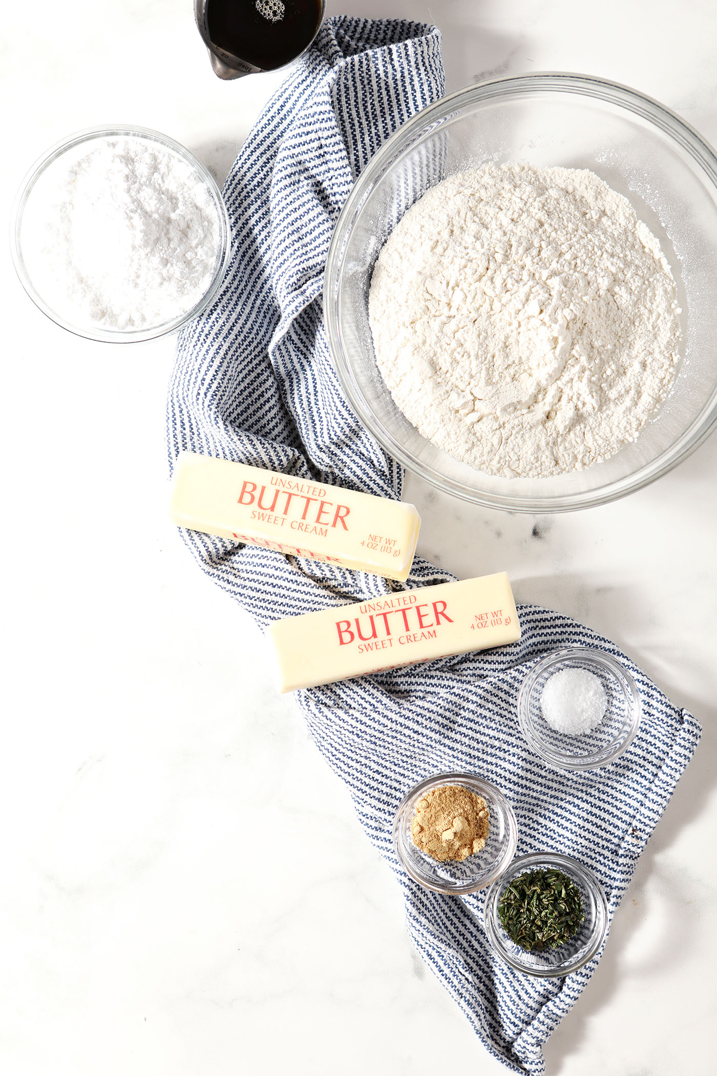 Ingredients to make ginger rosemary shortbread in bowls on a white stone surface with a blue and white striped linen