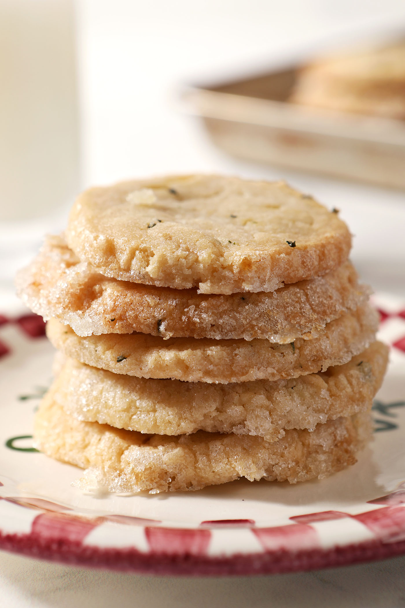 A stack of rosemary ginger shortbread cookies on a Christmas plate in front of a sheet pan and container of milk
