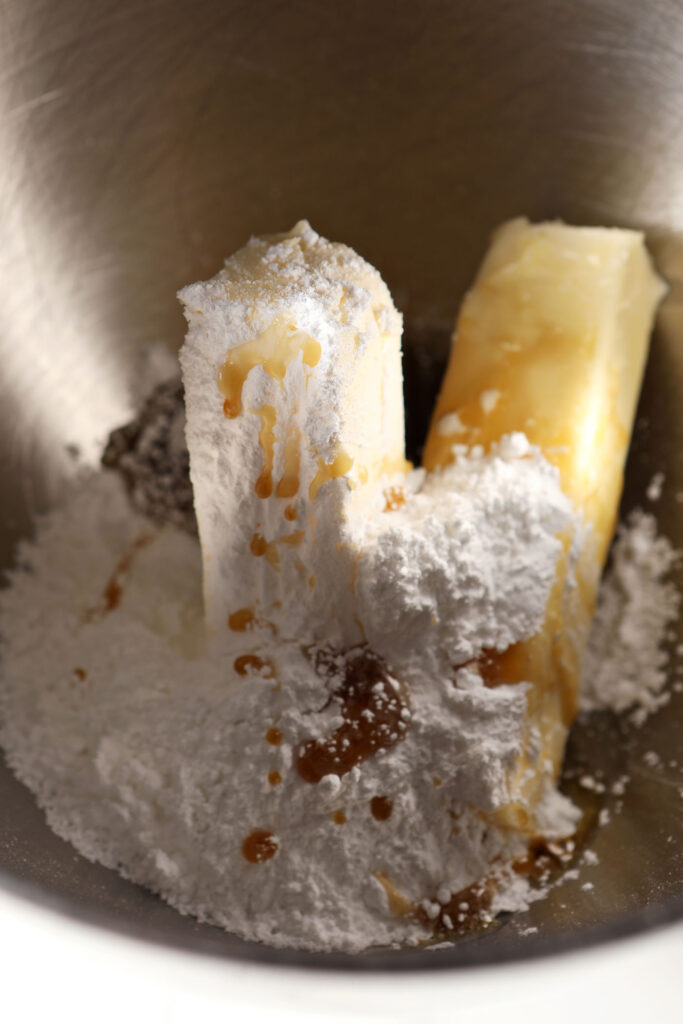 Two sticks of butter with powdered sugar and maple syrup in the metal bowl of a stand mixer before blending