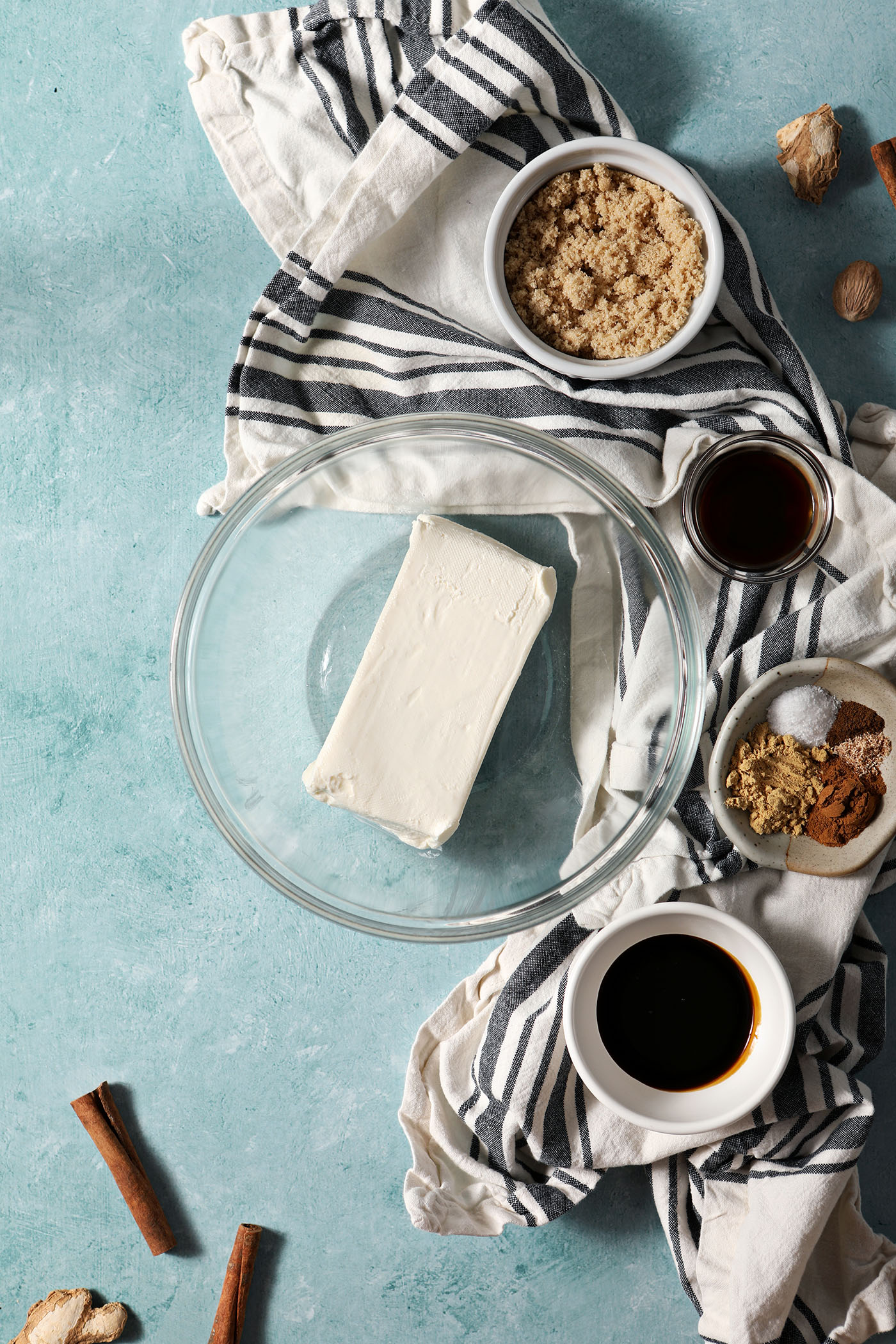 A bowl of cream cheese surrounded by other ingredients in bowls to make a holiday cookie dip on a teal background with a black and white striped linen