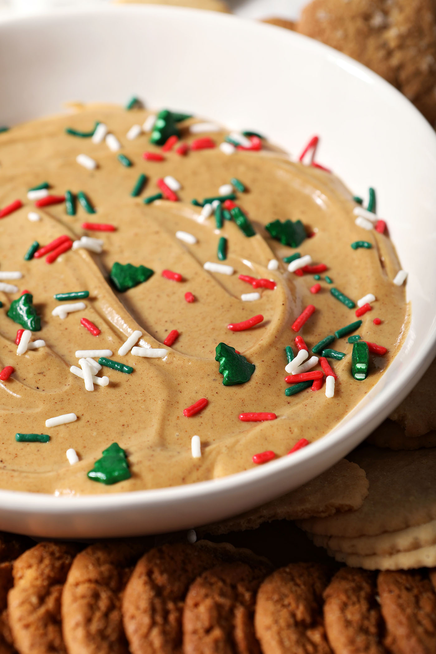 Closeup of gingerbread cheesecake dip in a white bowl garnished with Christmas sprinkles and surrounded by cookies