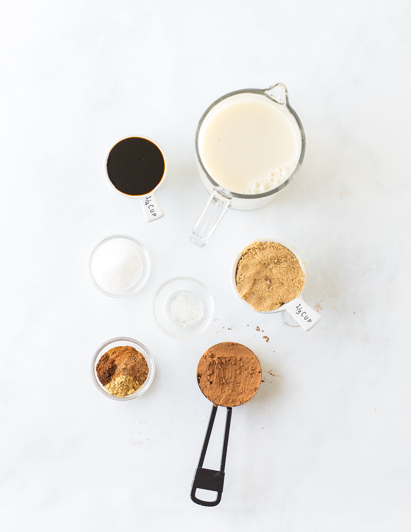 Ingredients to make gingerbread hot cocoa in cups and mugs on a white surface