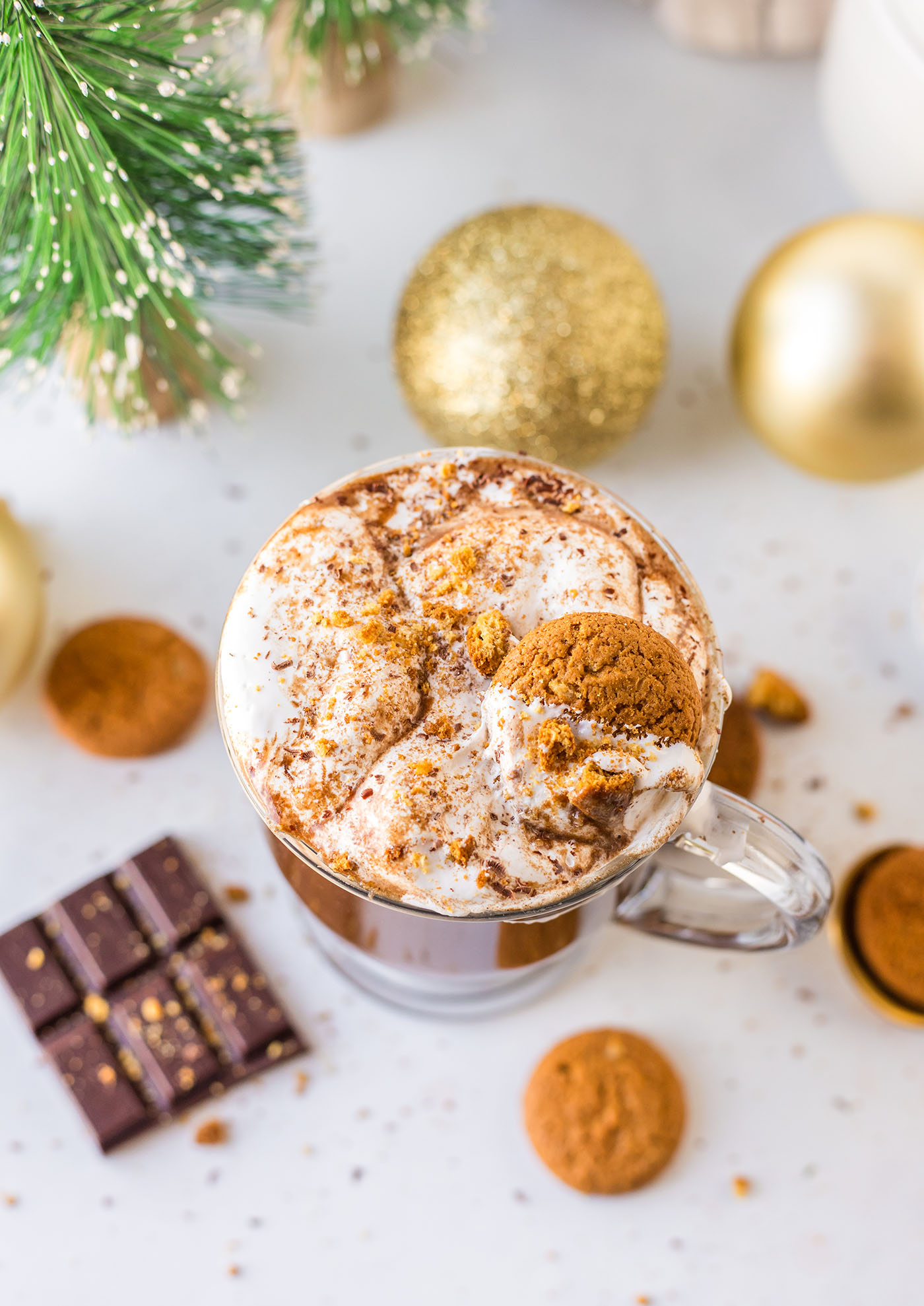 Gingerbread hot chocolate surrounded by chocolate, gingersnaps, ornaments and little Christmas trees on a white surface