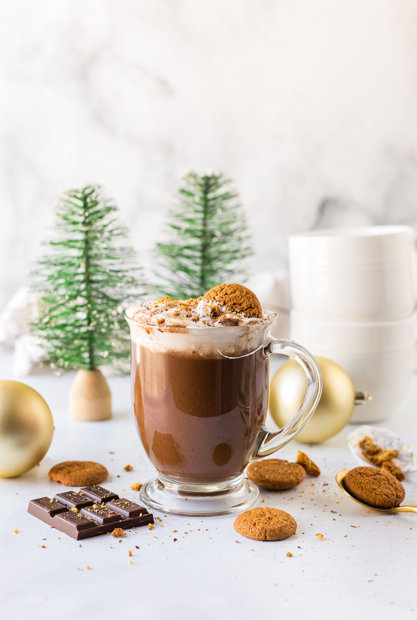 A mug of gingersnap hot chocolate on a white surface surrounded by chocolate, gingersnaps, ornaments and little trees