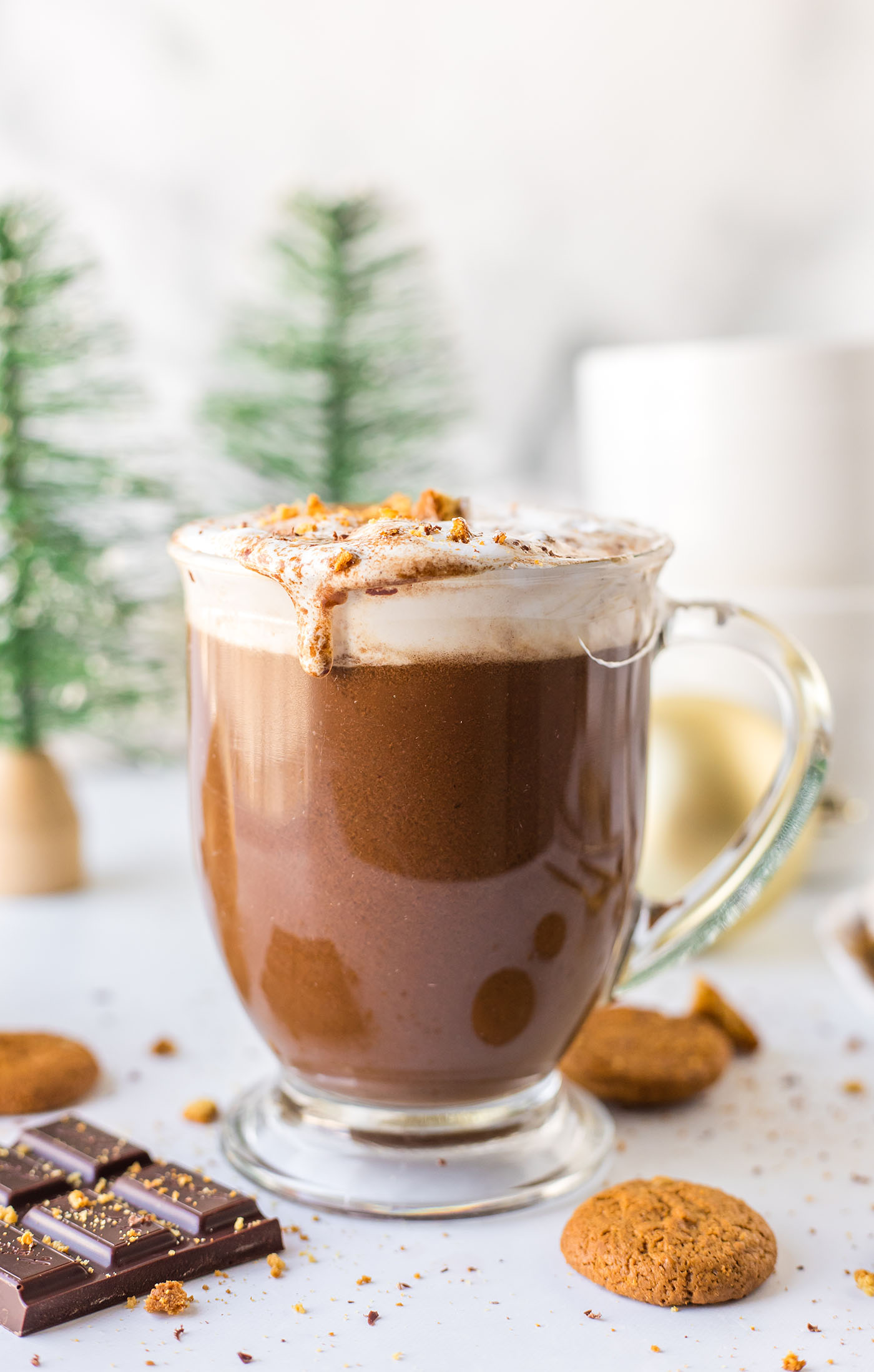 Closeup of a mug of gingerbread hot chocolate on a white surface surrounded by Christmas decor and ingredients