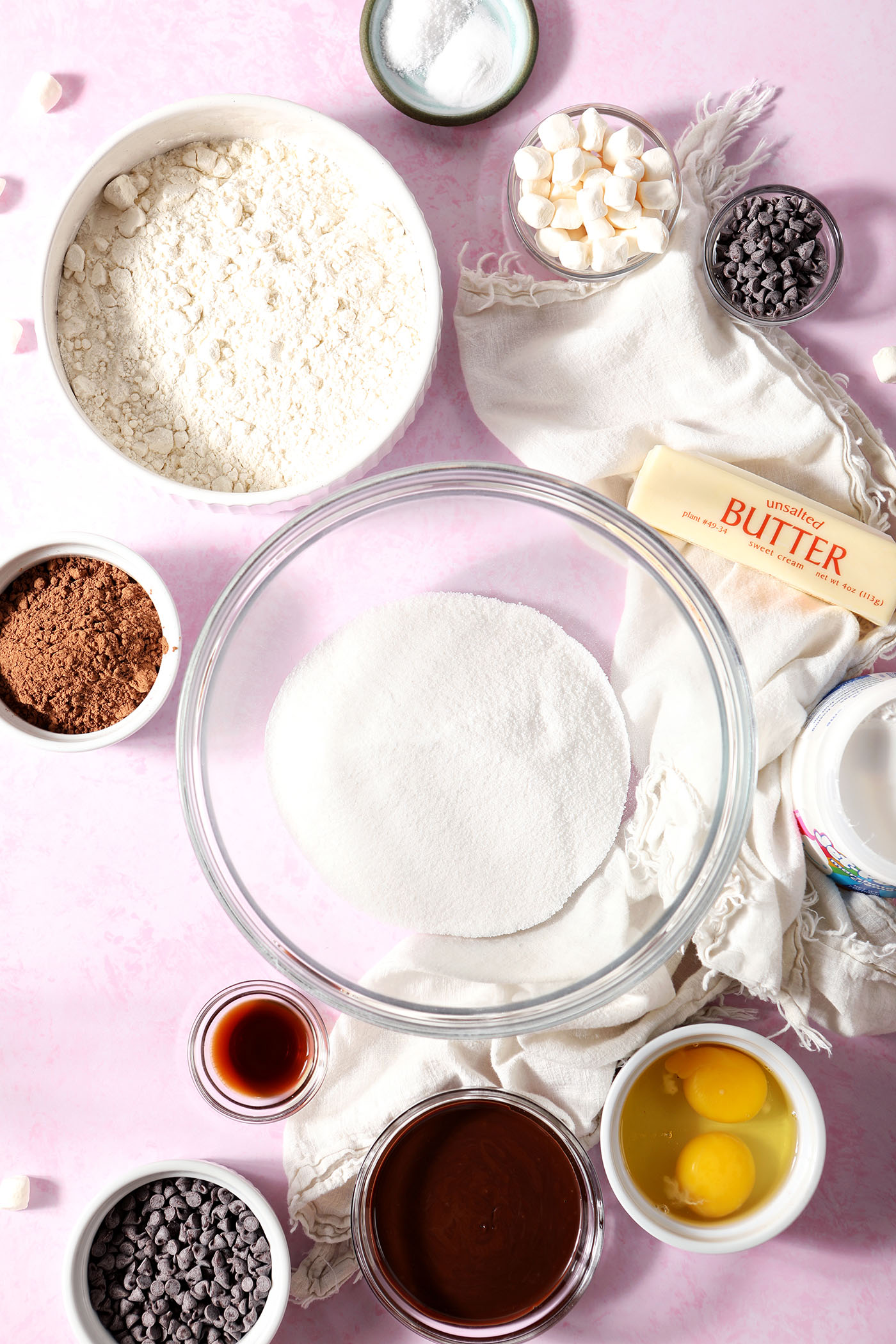 Bowls of ingredients to make hot chocolate cookies on a pink surface next to white linens