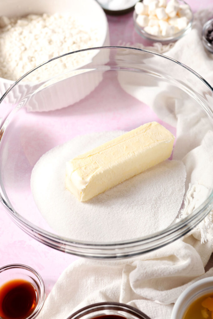 A stick of butter and granulated sugar in a glass bowl on a pink surface surrounded by other bowls