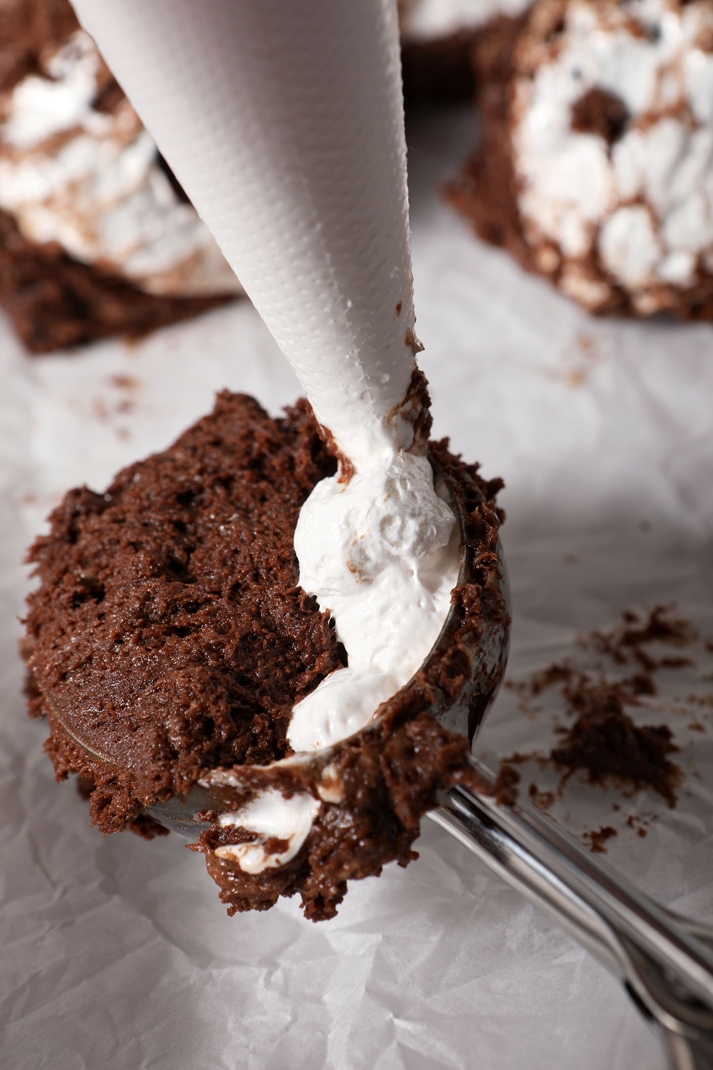 Marshmallow fluff is piped into a metal cookie scooper of cookie dough on a parchment-lined surface