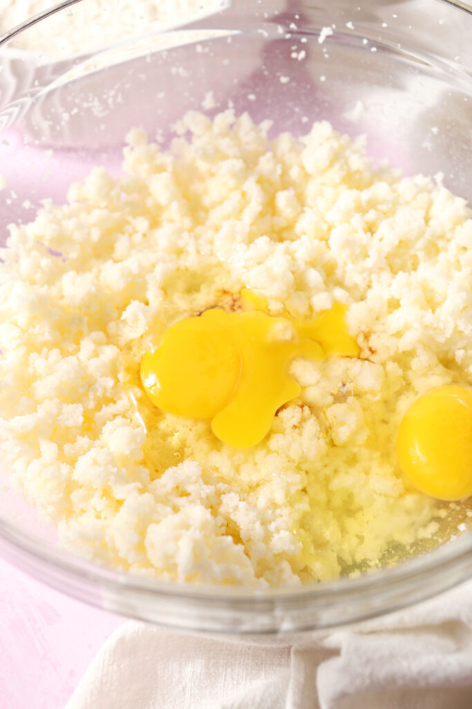 Eggs and vanilla sit on top of creamed butter and sugar in a bowl on a pink surface
