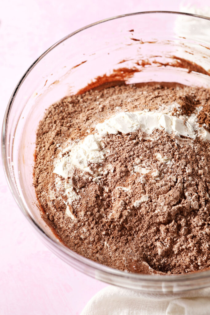 Dry ingredients sit on top of wet ingredients in a bowl on a pink surface before mixing